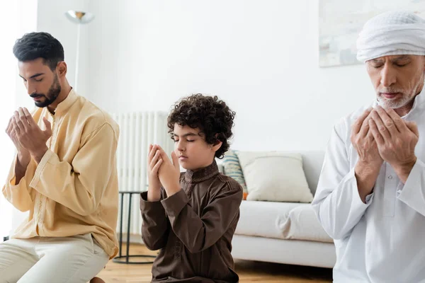 Preadolescent arabian kid with closed eyes praying near multiethnic men at home — Stock Photo