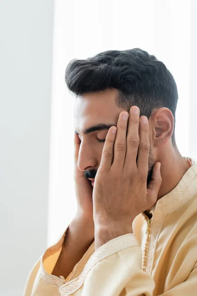 Hombre joven musulmán tocando la cara mientras reza en casa - foto de stock