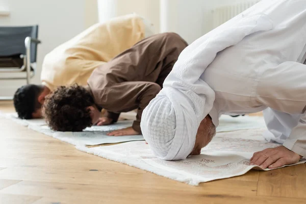 Uomo maturo piegandosi sul tappeto mentre prega vicino alla famiglia araba a casa — Foto stock