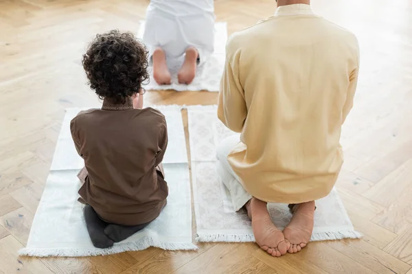 Vista trasera del niño musulmán rezando cerca de papá y abuelo en casa - foto de stock