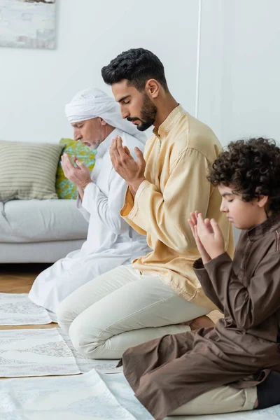 Side view of young muslim man praying near father and son on rugs at home — Stock Photo