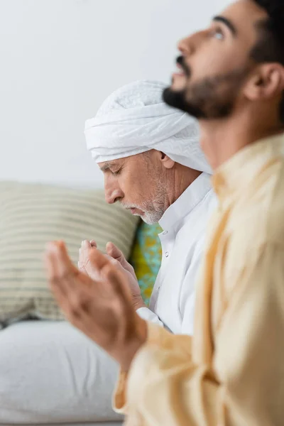 Side view of mature man praying with closed eyes near blurred arabian son at home — Stock Photo