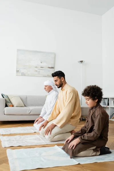 Hommes interraciaux et garçon préadolescent assis sur des tapis tout en priant à la maison — Photo de stock