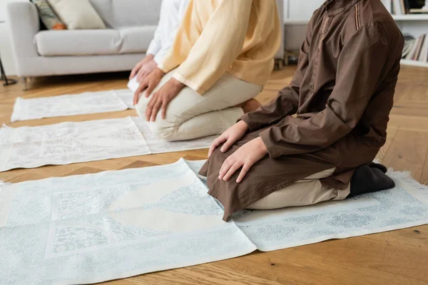 Vista ritagliata del ragazzo musulmano preadolescente seduto sul tappeto tradizionale mentre prega con la famiglia a casa — Foto stock