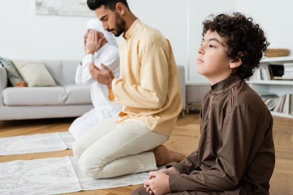 Curly menino muçulmano rezando perto de pai borrado e avô em tapetes em casa — Fotografia de Stock