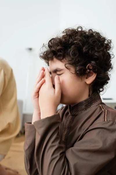 Lockiger arabischer Junge hält die Hände vor dem Gesicht, während er zu Hause in der Nähe des verschwommenen Vaters betet — Stockfoto