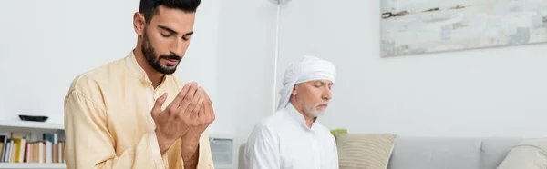 Young arabian man in traditional clothes praying near mature dad at home, banner — Stock Photo