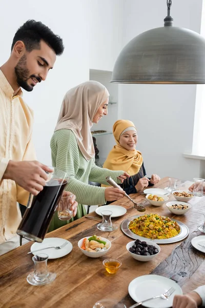 Mujer árabe sonriente de pie cerca de la comida, marido con té y mamá asiática en casa - foto de stock
