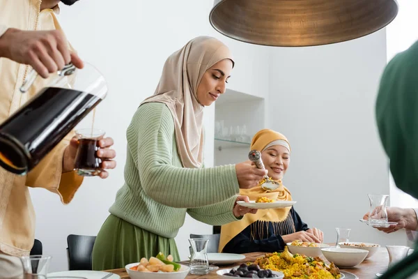 Mujer árabe sirviendo pilaf mientras marido vertiendo té y familia interracial en casa - foto de stock