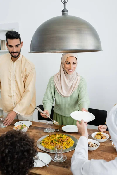 Mujer musulmana sonriente sirviendo pilaf cerca de la familia y la comida en casa - foto de stock