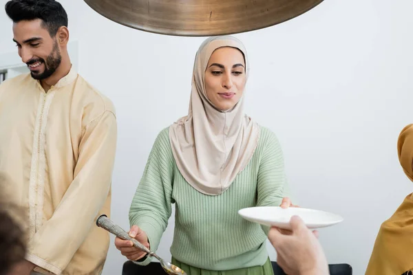 Mujer musulmana joven sosteniendo cuchara y plato cerca de la familia en casa - foto de stock
