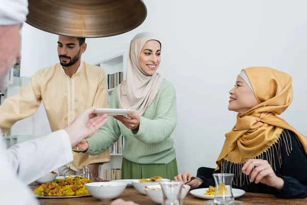 Feliz mujer musulmana sosteniendo plato cerca de pilaf y la familia interracial en casa - foto de stock