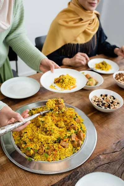 Vista recortada de la joven musulmana sirviendo pilaf cerca borrosa mamá y comida en casa - foto de stock