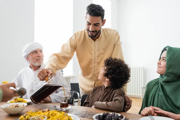 Glücklicher muslimischer Mann schenkt Tee in der Nähe einer interrassischen Familie und zu Hause Essen ein — Stockfoto