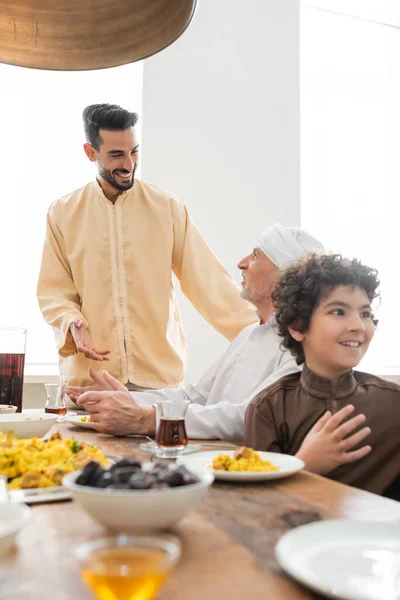 Lächelnder muslimischer Mann spricht mit reifem Vater in der Nähe von leckerem Essen zu Hause — Stockfoto