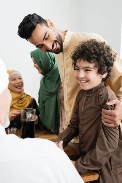 Sonriente hombre árabe abrazando hijo cerca del té y la familia multiétnica en casa - foto de stock