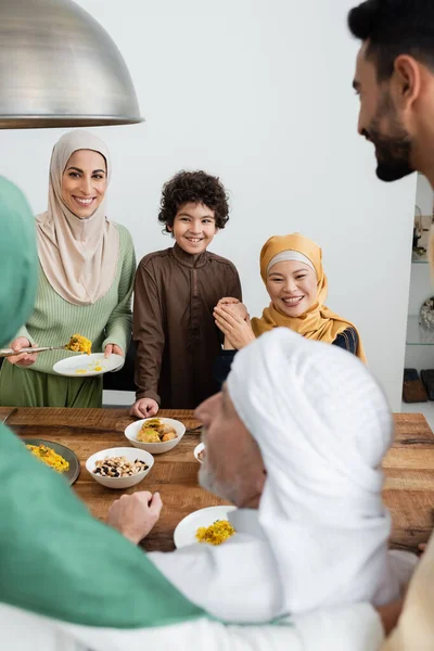 Família muçulmana multicultural sorrindo perto de comida na mesa em casa — Fotografia de Stock