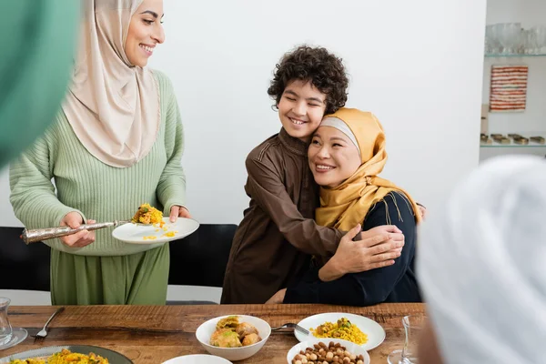 Sonriente musulmán chico abrazando asiático abuela cerca mamá y comida en casa - foto de stock