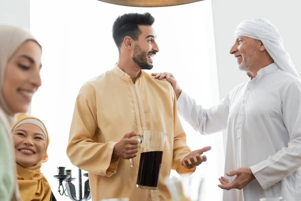 Aufgeregter arabischer Mann hält eine Kanne Tee in der Hand, während er mit seinem Vater in der Nähe einer interrassischen Familie spricht — Stockfoto