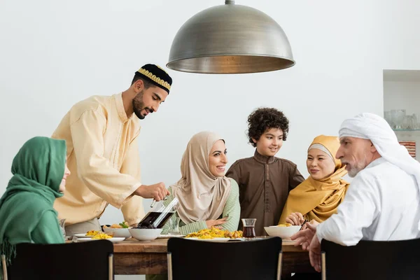 Young arabian man in skullcap pouring tea during dinner with interracial muslim family — Stock Photo
