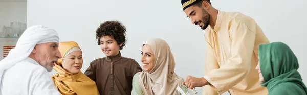 Familia musulmana multiétnica sonriendo cerca del hombre árabe con jarra en casa, pancarta - foto de stock
