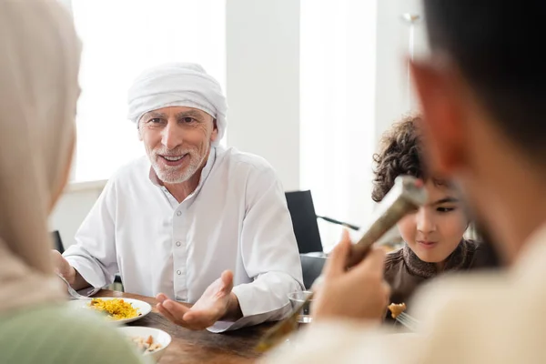 Uomo musulmano sorridente che punta con mano durante la cena con nipote arabo e famiglia offuscata — Foto stock
