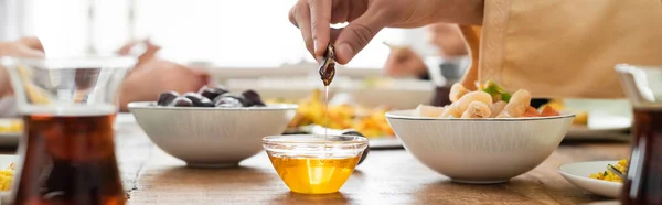 Cropped view of muslim man dipping date into honey near baklava and blurred tea, banner — Stock Photo