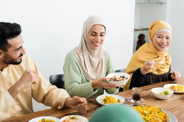 Pleased arabian woman in hijab holding bowl with nuts near asian mother and husband — Stock Photo