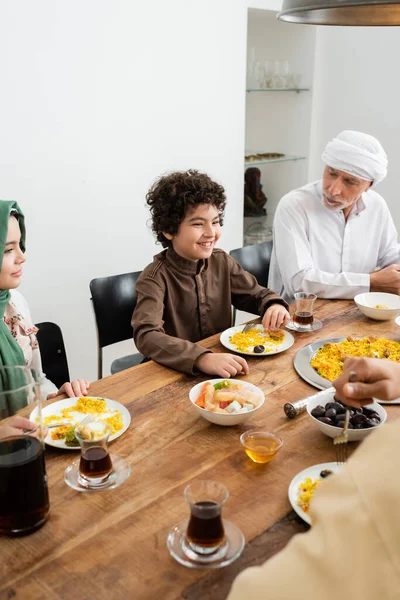Niños multiétnicos felices cenando cerca del abuelo musulmán en casa - foto de stock