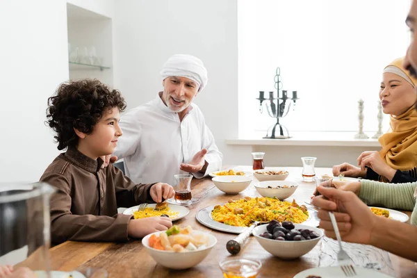 Homem muçulmano de meia idade apontando para o neto árabe durante o jantar de família com a família interracial — Fotografia de Stock