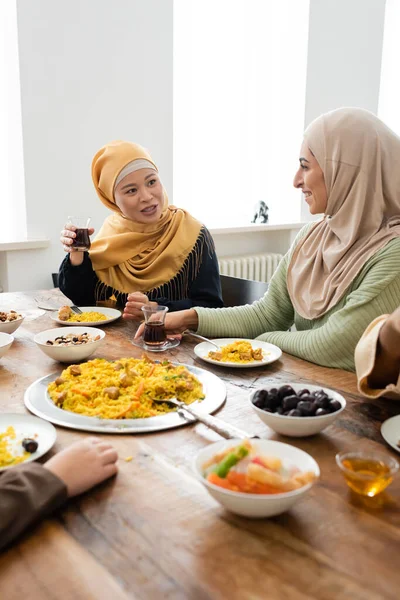 Asian muslim woman holding glass of tea near arabian daughter during family dinner — Stock Photo
