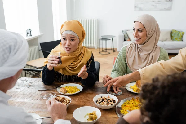 Asian woman pointing with hand while talking near smiling muslim daughter and blurred family — Stock Photo