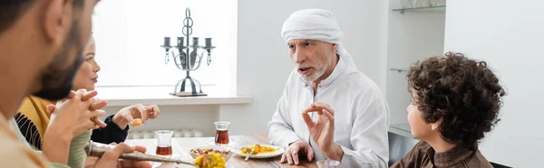 Mature muslim man gesturing while talking during dinner with multicultural family, banner — Stock Photo