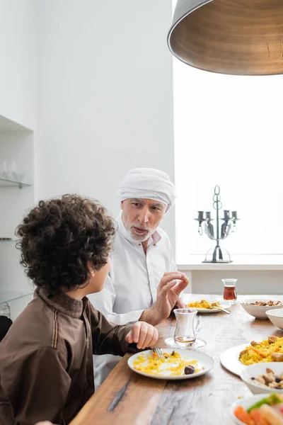 Maduro musulmán hombre gesto mientras habla con el nieto árabe durante la cena familiar - foto de stock