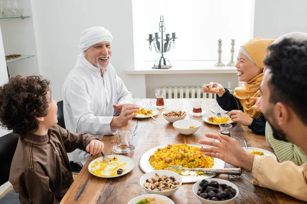 Feliz homem muçulmano de meia idade falando com a família interracial durante o jantar em casa — Fotografia de Stock