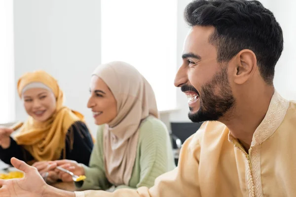 Cheerful muslim man pointing with hand near interracial women eating pilaf for dinner — Stock Photo