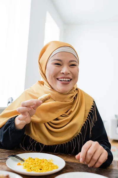 Cheerful asian woman in hijab holding sweet baklava near pilaf on plate — Stock Photo