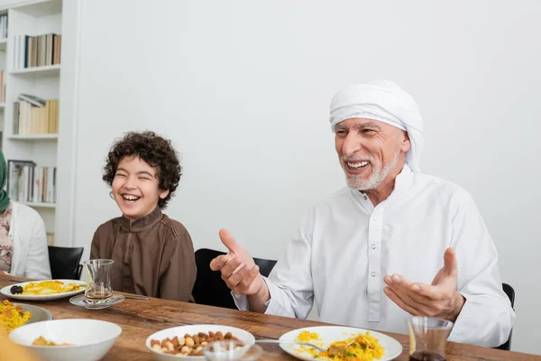 Happy muslim man pointing with hands near laughing arabian grandson during family dinner — Stock Photo