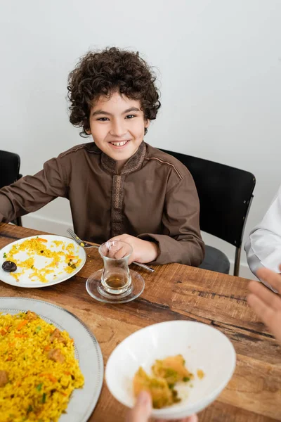 Счастливый арабский мальчик-мусульманин, улыбающийся во время ужина с размытой семьей — стоковое фото