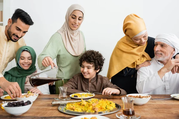 Araberin schenkt Tee in der Nähe einer glücklichen multiethnischen muslimischen Familie ein, die zu Hause zu Abend isst — Stockfoto