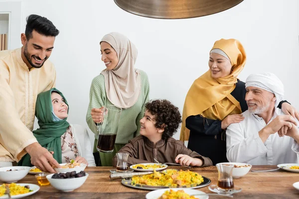 Mujer árabe joven sosteniendo jarra de té cerca de la familia musulmana multiétnica cenando en casa - foto de stock