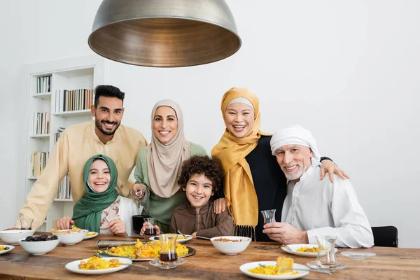 Happy multiethnic muslim family looking at camera near pilaf and tea on table — Stock Photo