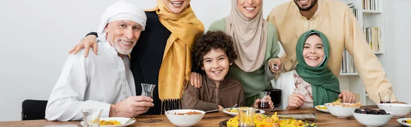 Familia musulmana multiétnica sonriendo a la cámara cerca de pilaf y té en la mesa, pancarta - foto de stock