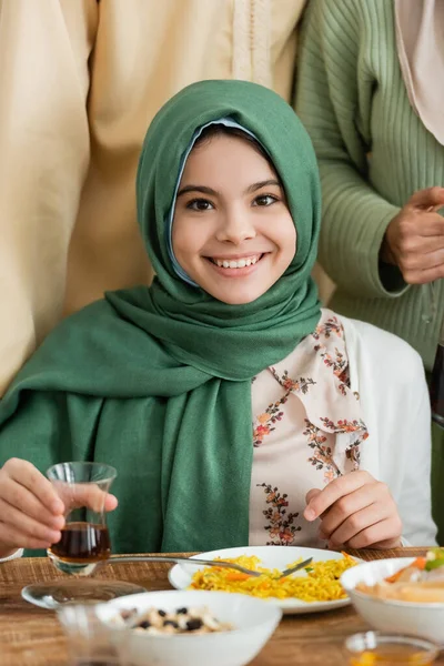 Menina muçulmana feliz no hijab olhando para a câmera enquanto segurando um copo de chá perto dos pais — Fotografia de Stock