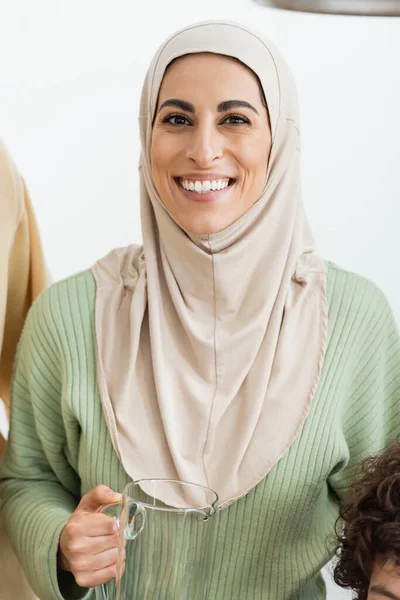 Glücklich muslimische Frau im Hijab hält Krug, während sie in die Kamera schaut — Stockfoto