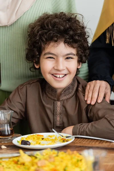 Alegre musulmán árabe chico mirando la cámara cerca de pilaf durante la cena familiar - foto de stock