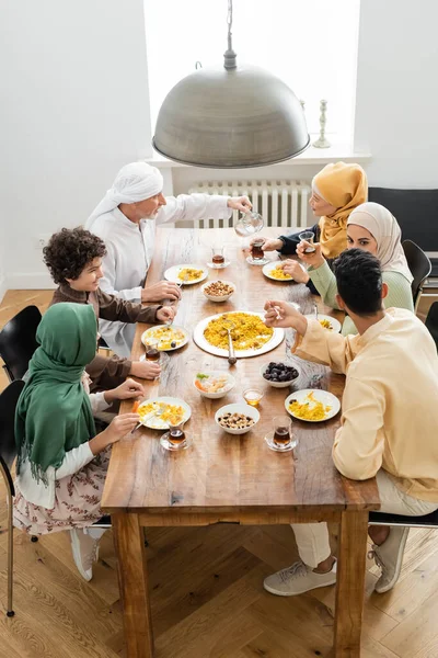 Vista de ángulo alto del hombre musulmán de mediana edad sirviendo té durante la cena con la familia multiétnica - foto de stock