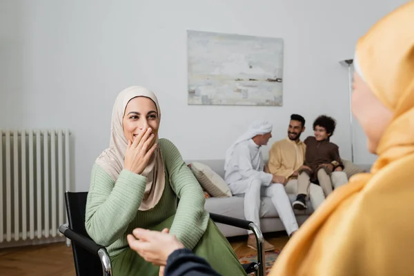 Riéndose mujer musulmana cubriendo la boca con la mano cerca de la familia multiétnica sobre fondo borroso - foto de stock