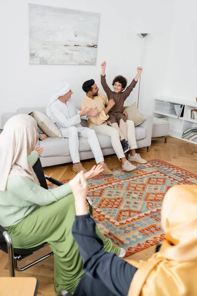 Возбужденный арабский мальчик показывает победу жест возле межрасовой мусульманской семьи в гостиной — стоковое фото