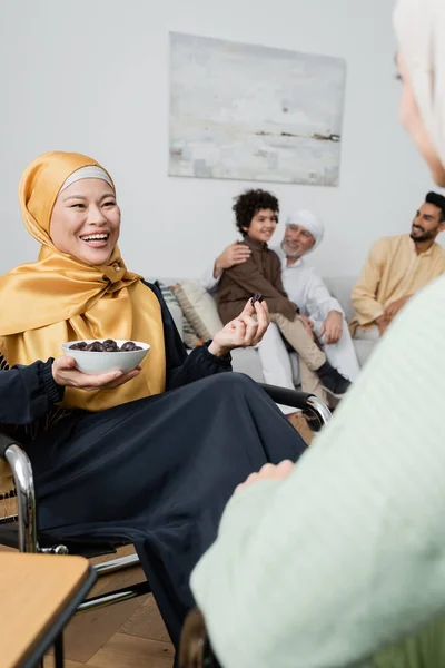 Glücklich asiatische Frau im Hijab hält Schüssel mit Daten in der Nähe verschwommene Tochter und multiethnische muslimische Familie — Stockfoto